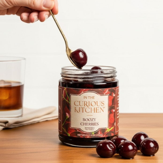 A decorative gold spoon lifting a bourbon-infused cocktail cherry from a jar, next to a glass of whiskey on a wooden table.