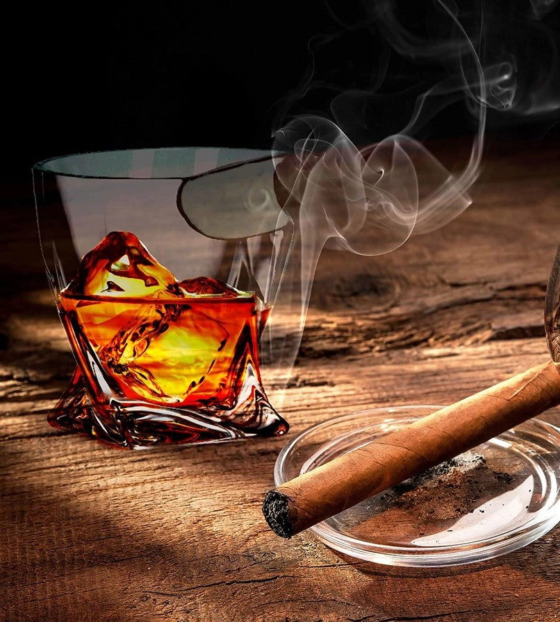 A single Cigar Whiskey Glass in the modern Twist style with ice and a smoking cigar on a wooden surface