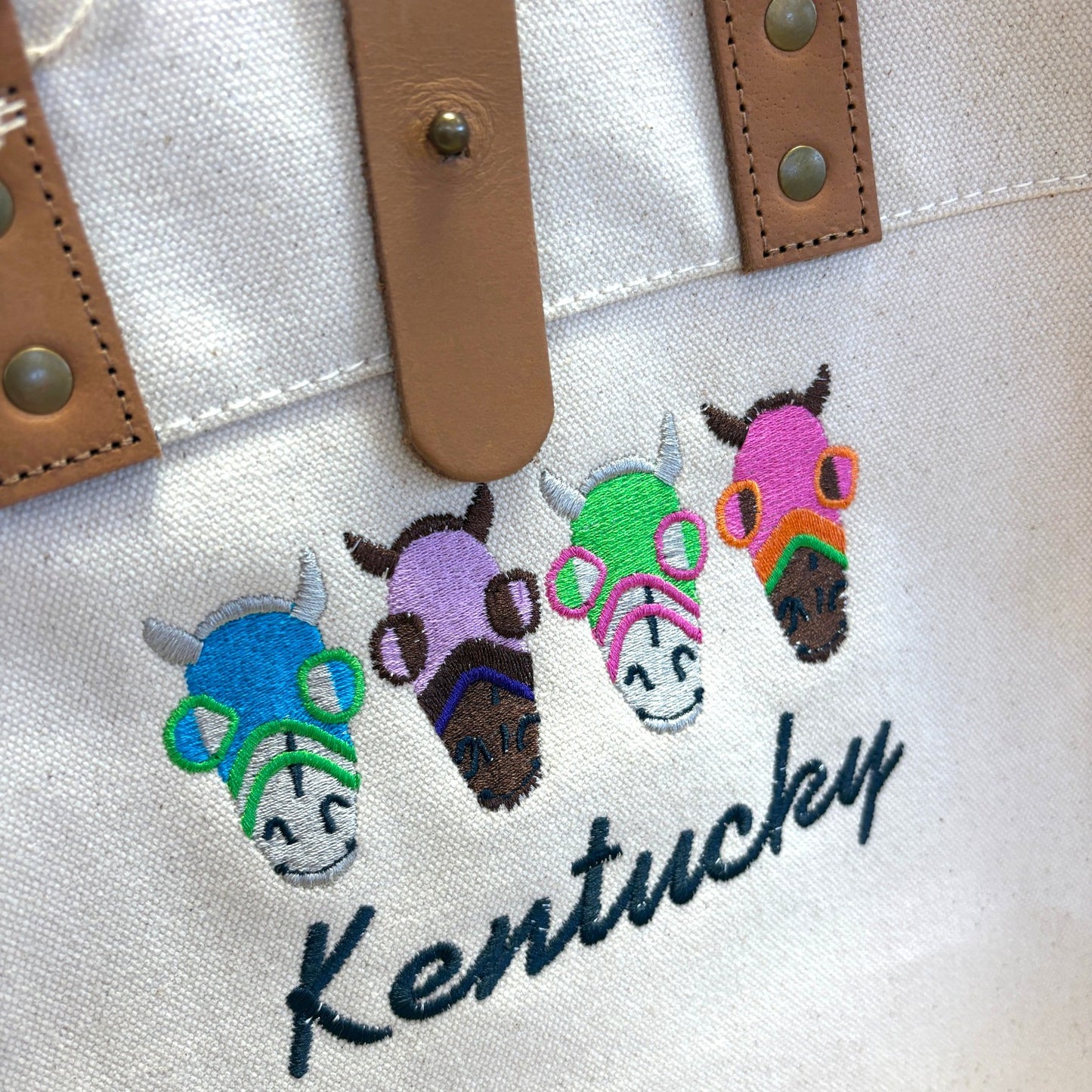 Close-up of the vibrant Kentucky Derby embroidery showing four stylized horses wearing blue, purple, green, and pink racing hoods.