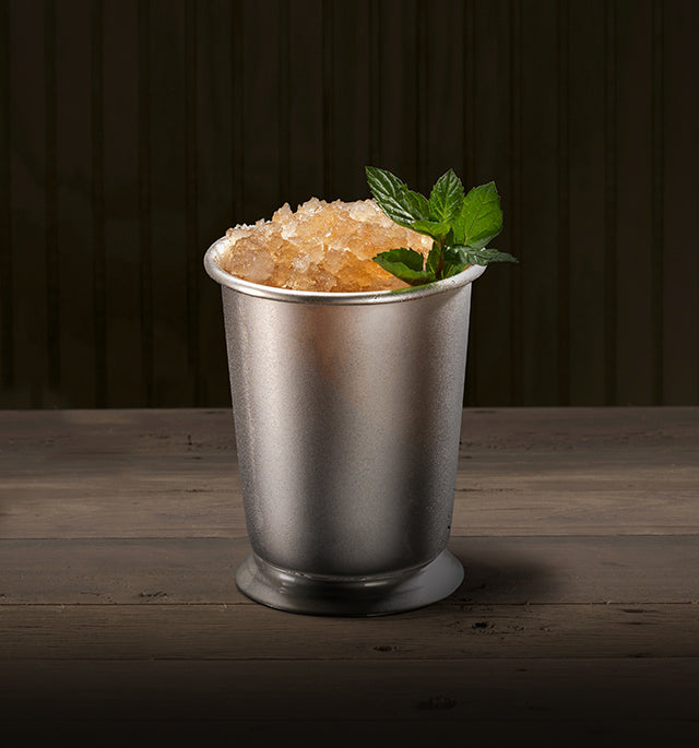 Metallic julep cup with crushed ice and mint leaves on a wooden surface with a dark background