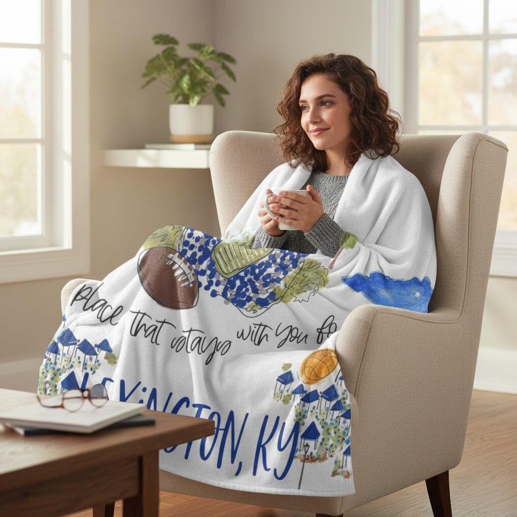 Woman sitting in a cream armchair, wrapped in a Lexington KY themed fleece blanket with hand-drawn artwork and sports motifs.