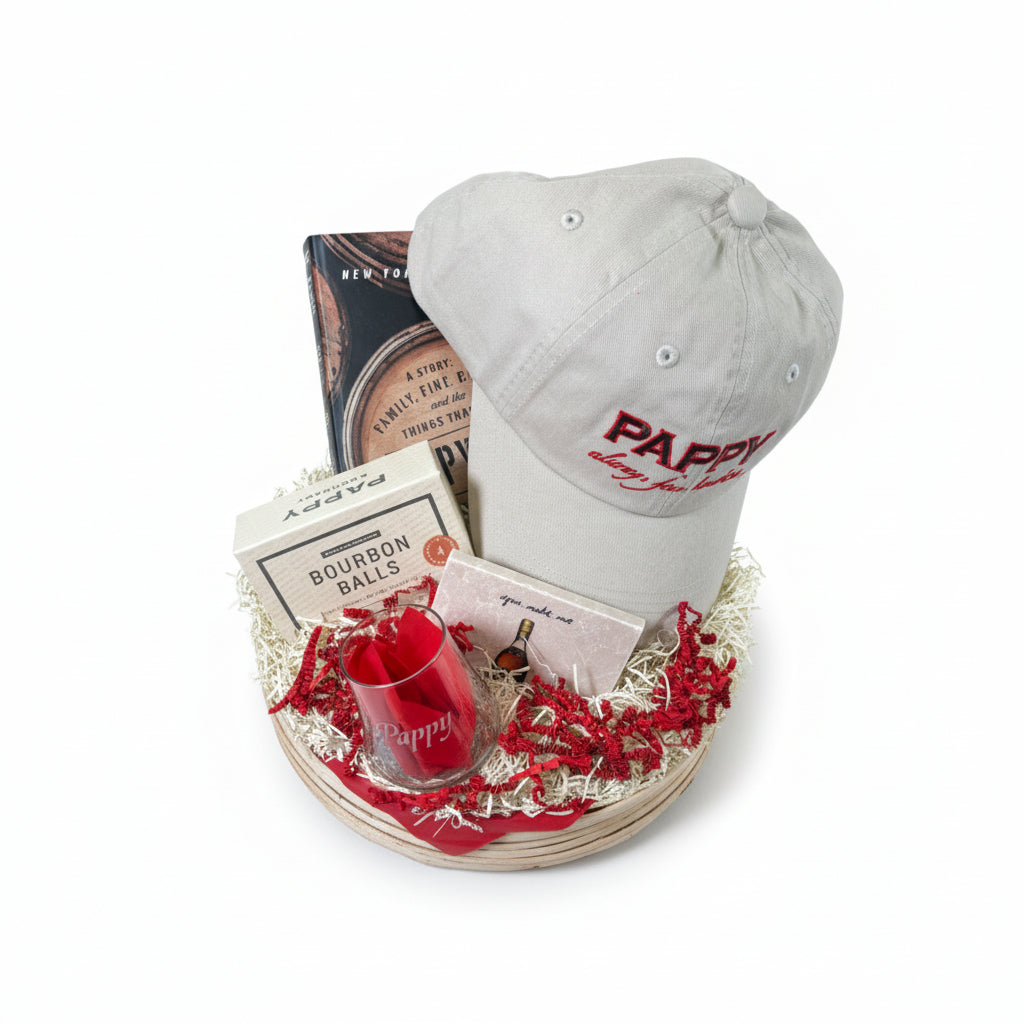Gray cap with 'Pappy' branding, bourbon balls, and a book in a decorative basket on a white background