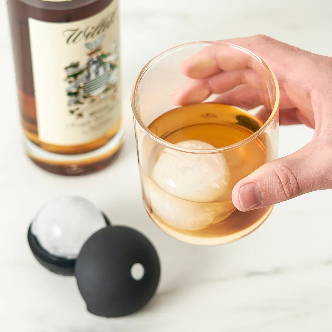 Hand holding a glass of whiskey with spherical ice cubes, next to a bottle of whiskey and two silicone ice cube molds.