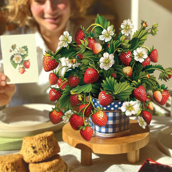 Decorative strawberry plant on a wooden stand with a person holding a card in the background.