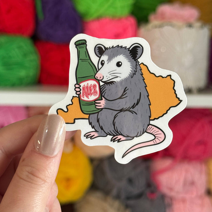 Sticker of an opossum holding a bottle of ale-8 with yarn balls in the background