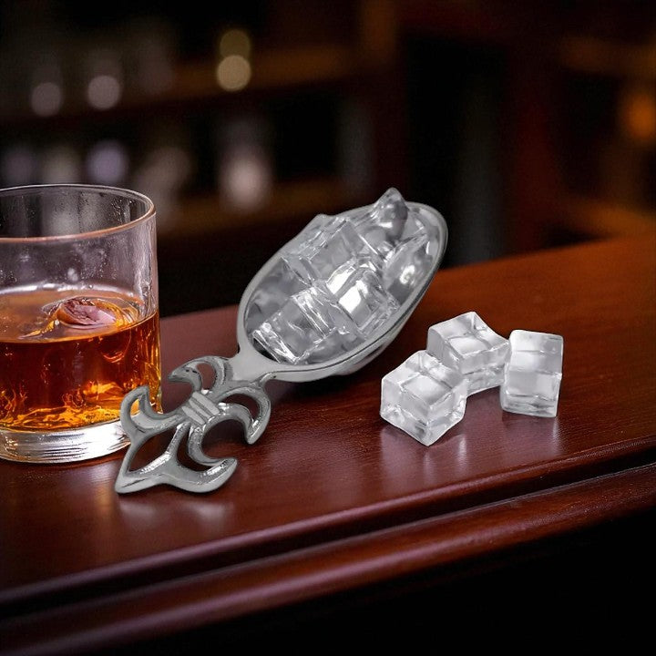 Silver ice scoop with ice cubes and a glass of whiskey on a wooden surface.