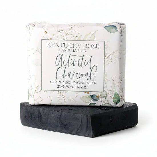 Handcrafted activated charcoal hemp facial soap bar wrapped in botanical paper sitting on top of an unwrapped black soap bar against a white background.
