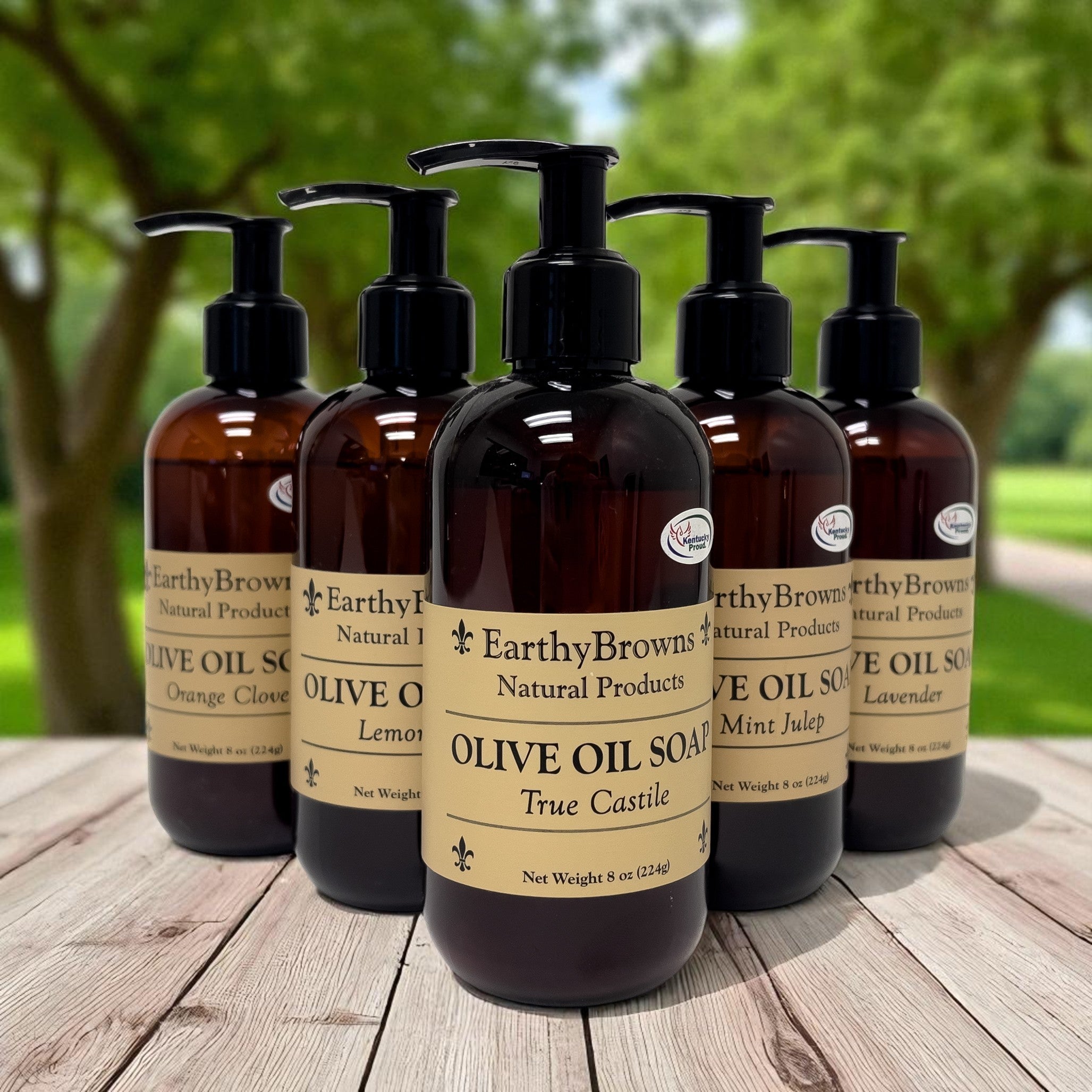 Five bottles of Earthy Browns Natural Products olive oil soap on a wooden surface with a natural background.