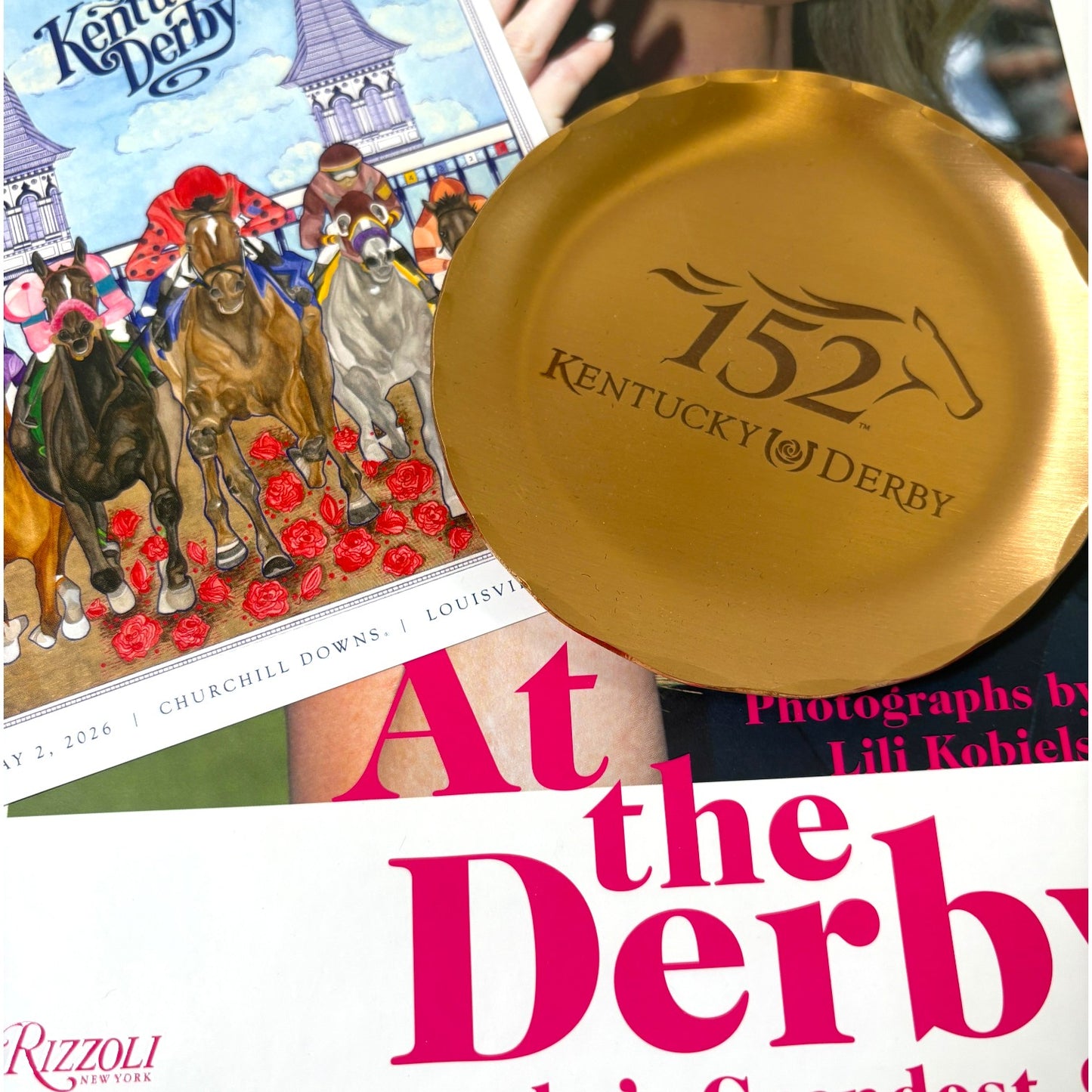 Close up of the Kentucky Derby 152 Keepsake Bundle featuring the At the Derby Rizzoli coffee table book, official 152nd bronze coaster, and Art of the Derby postcard.`