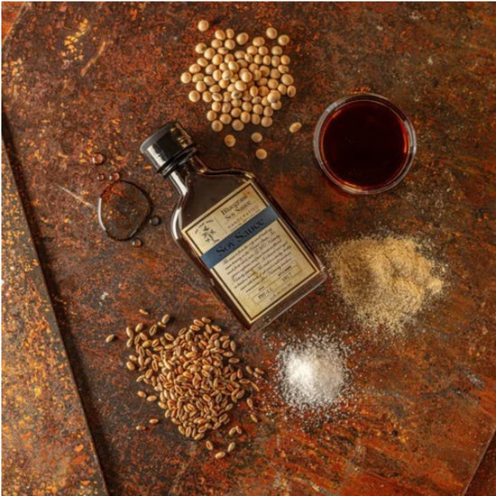 Bluegrass Soy Sauce bottle surrounded by soybeans, wheat, sea salt, and soy sauce on a textured surface