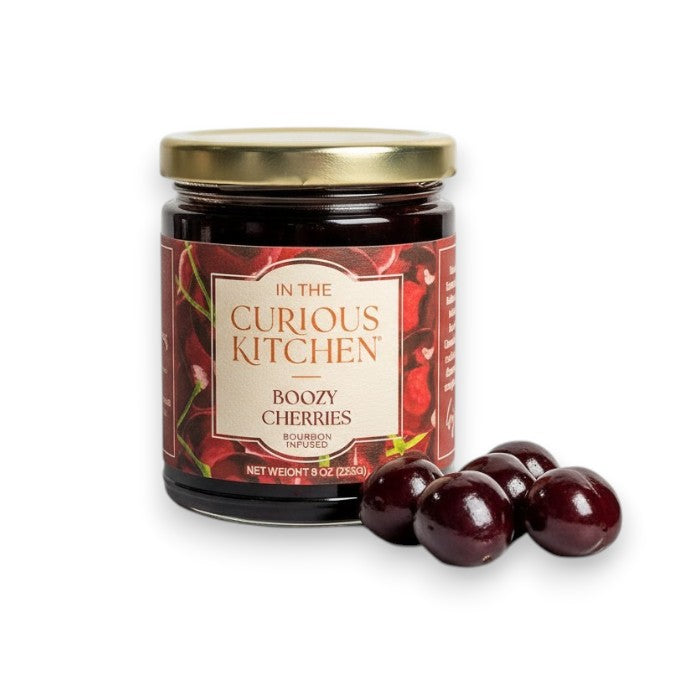 A jar of Boozy Cherries with a cream-colored label surrounded by five fresh, dark red cherries on a white background.