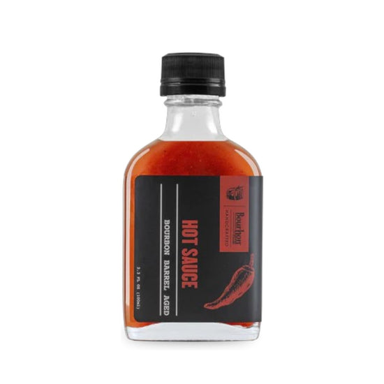 A glass bottle of Bourbon Barrel Aged Hot Sauce with a black cap, set against a rustic background.