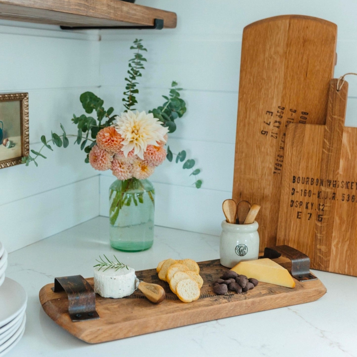 A styled kitchen scene with wooden serving boards and charcuterie food on a white countertop. A glass jar with flowers sits in the background, and a large wooden cutting board with printed text stands upright behind the boards.