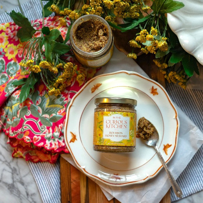 A jar of coarse-grain Bourbon Honey Mustard sitting on a white decorative plate next to a silver spoon holding a dollop of mustard, surrounded by floral accents.