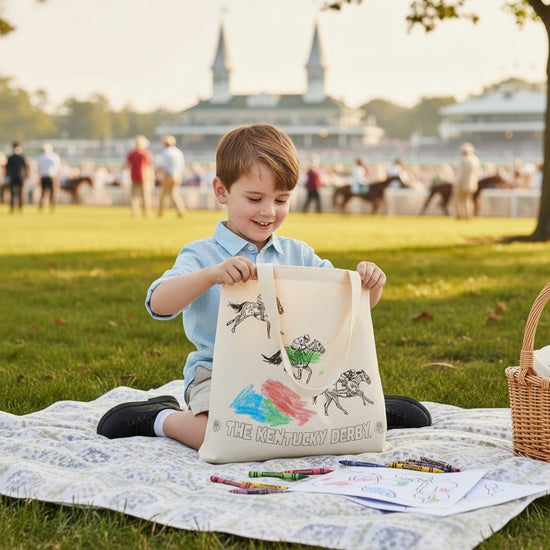 A young boy sits on a blanket with the iconic Churchill Downs spires in the background, happily coloring the Kentucky Derby tote bag.