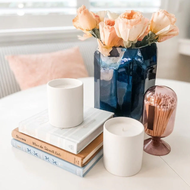Decorative setting with books, a white mug, a blue vase with peach flowers, and a pink candle holder on a table.