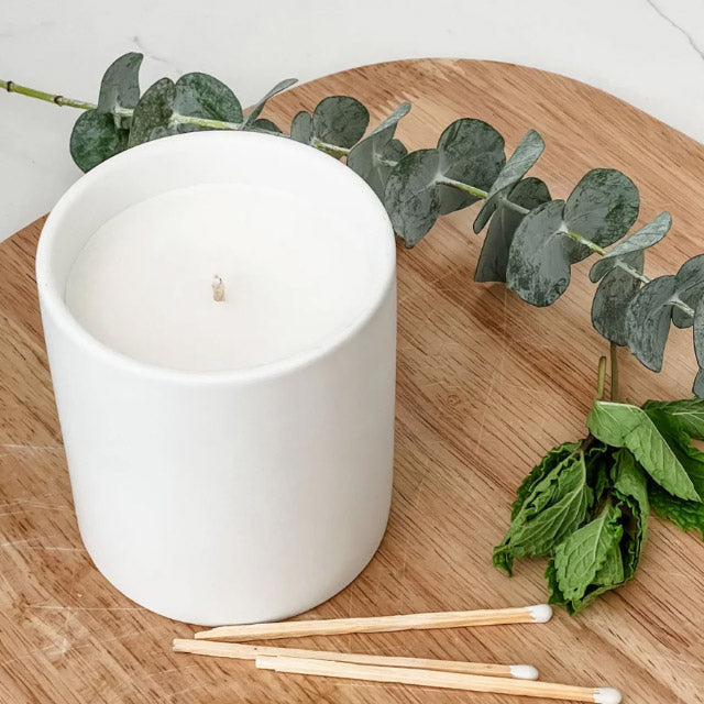 White candle in a white container on a wooden surface with eucalyptus leaves and matches.