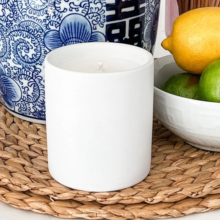 White candle on a woven mat with a blue and white patterned vase and bowl of fruit in the background.