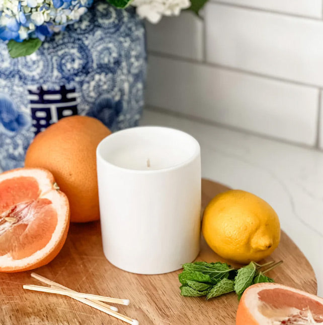 White candle on a wooden surface with lemons, oranges, and a floral arrangement in the background.
