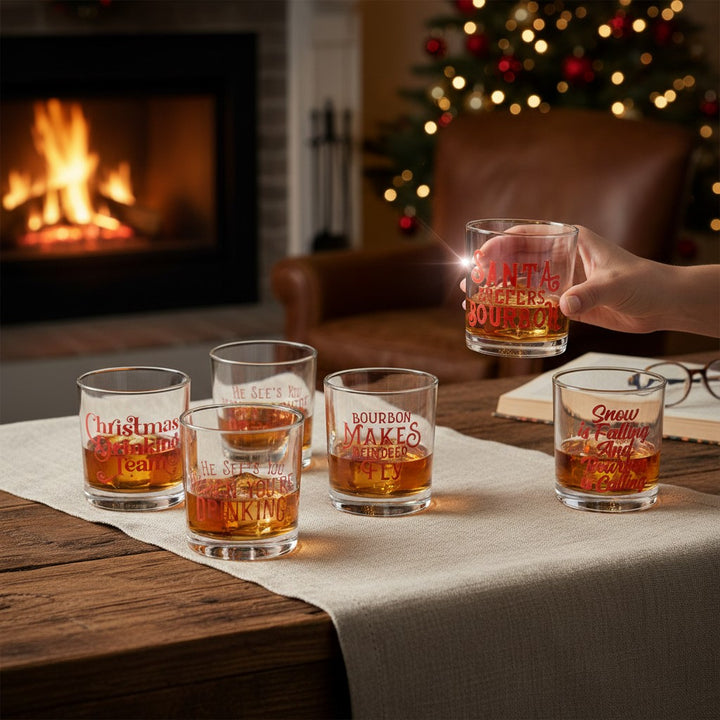 Set of six Christmas-themed festive rocks glass filled with bourbon and ice on a table with a fireplace and decorated tree in the background.