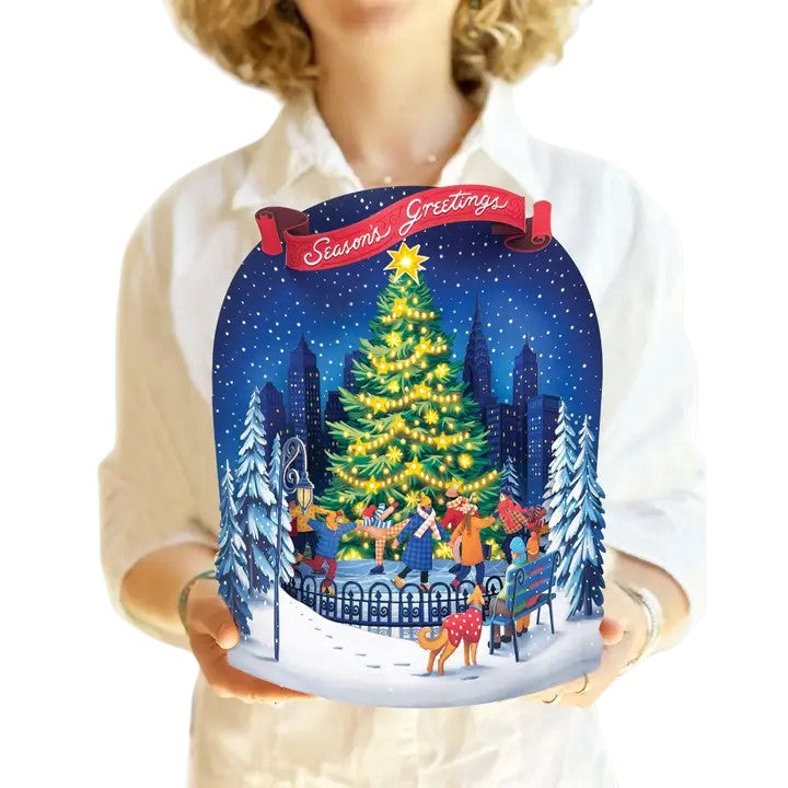Person holding a Christmas-themed decorative item with a tree and people around it.