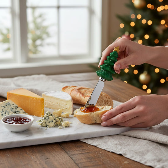 Person spreading jam on bread with a Christmas tree-shaped cheese spreader, surrounded by cheese and bread on a table with a festive background.