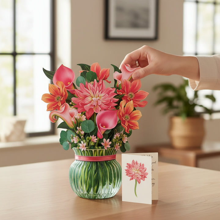 Hand placing a pink flower into The Dear Dahlia paper flower bouquet fully opened, featuring pink dahlias, calla lilies, and blue eucalyptus branches.