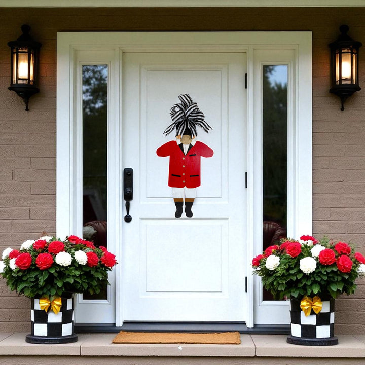 Decorative door with a person in a red coat and black hat, flanked by two flower pots with red and white flowers on a brick wall.