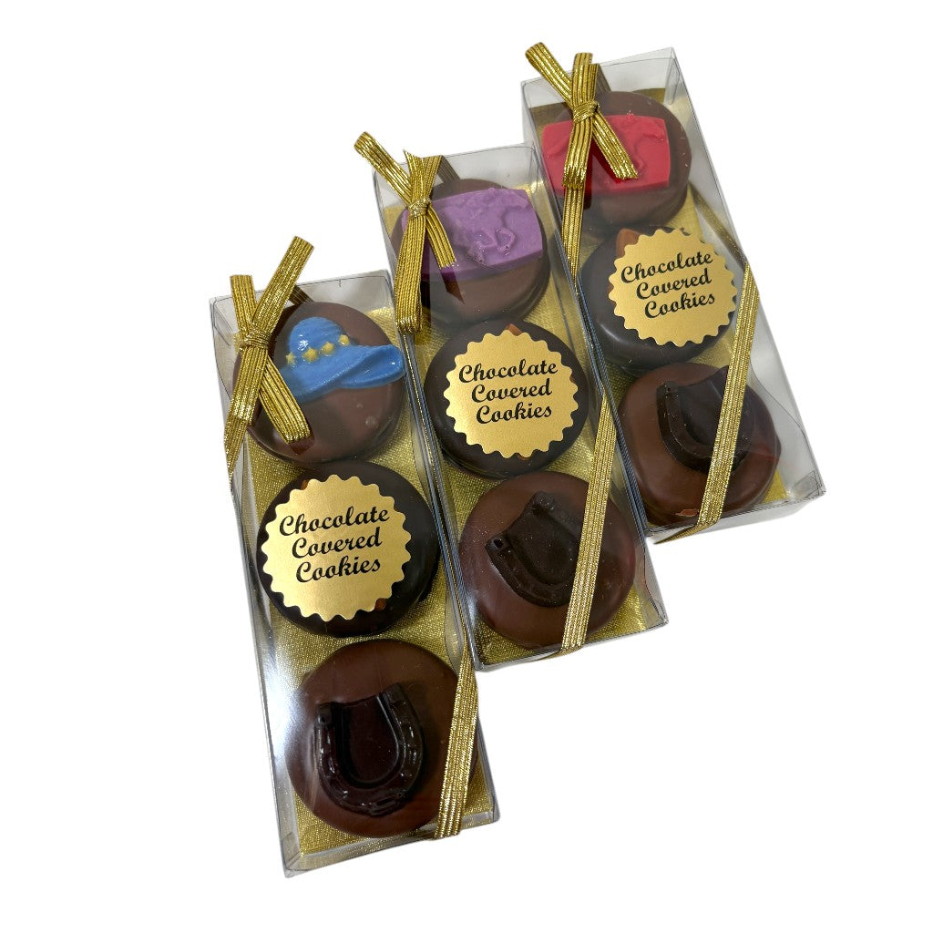 Clear gift boxes tied with shiny gold ribbons, containing decorated chocolate covered sandwich cookies resting on gold shred.