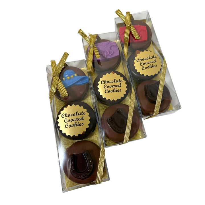 Clear gift boxes tied with shiny gold ribbons, containing decorated chocolate covered sandwich cookies resting on gold shred.