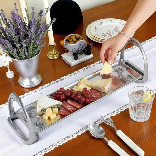 A beautifully arranged charcuterie board on a wooden table, featuring an assortment of cheeses, meats, and grapes displayed on the silver stirrup-handled tray.