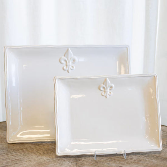 Two white ceramic trays with fleur-de-lis designs on a wooden surface.