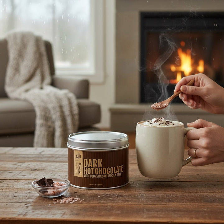 A lifestyle scene showing a mug of hot chocolate next to the Bourbon Barrel Foods tin by a cozy fireplace.