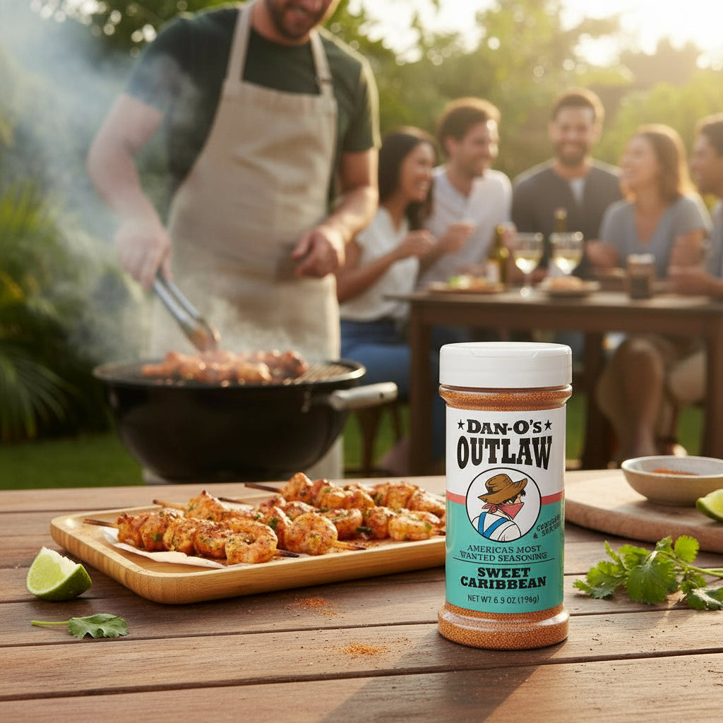 Bottle of Dan-O's Outlaw Sweet Caribbean seasoning on a wooden table with people grilling and dining outdoors.
