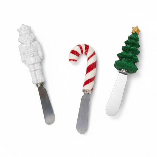 The three holiday cheese spreaders, a white nutcracker, a red-and-white candy cane, and a green Christmas tree, on a white background.