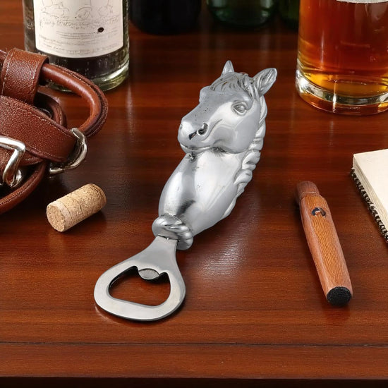 Silver horse-shaped bottle opener on a wooden surface with a glass of amber liquid and leather items.