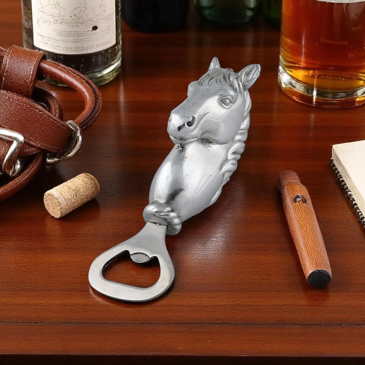 Silver horse-shaped bottle opener on a wooden surface with a glass of amber liquid and leather items.