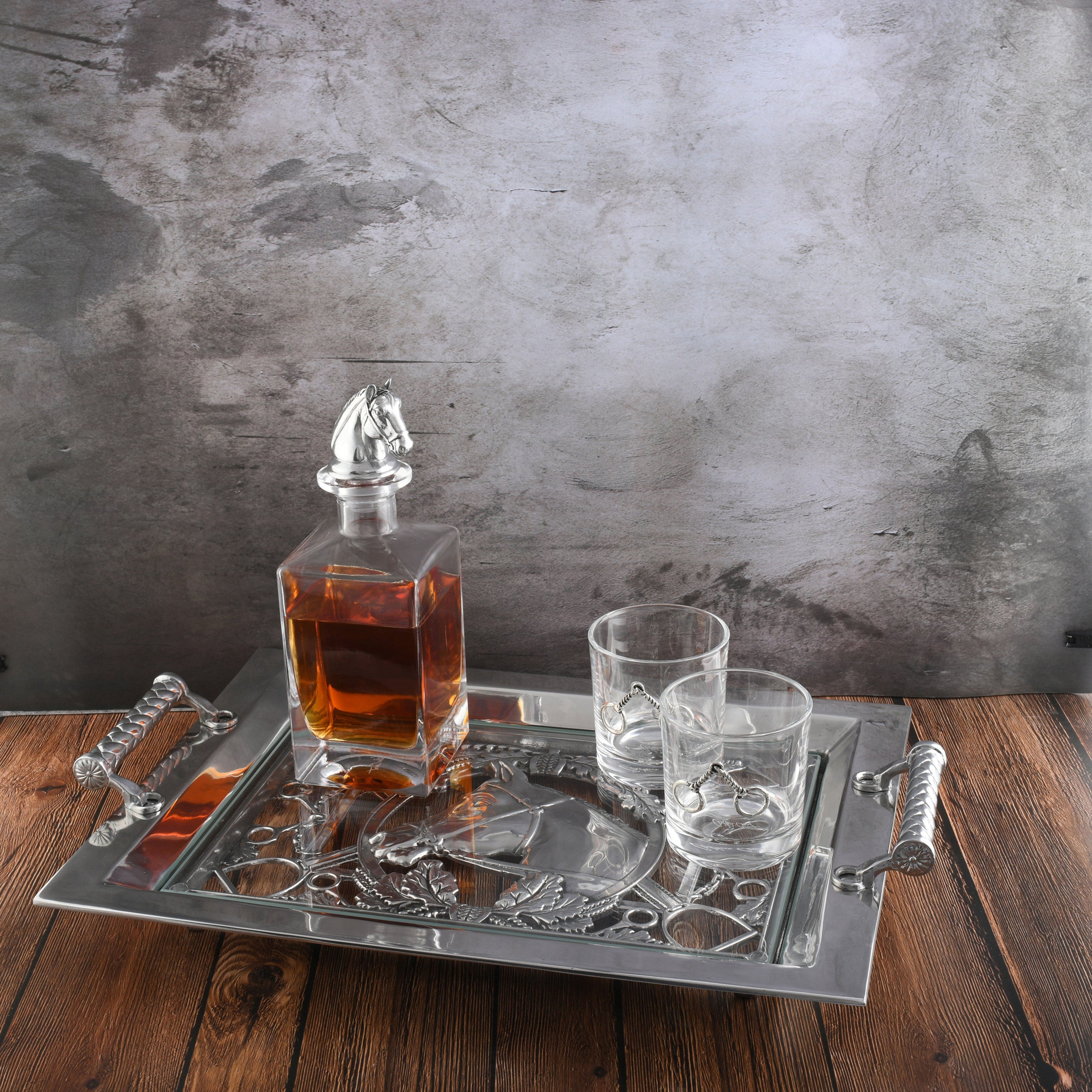 Alcohol decanter with horse head design on a decorative tray with glasses against a textured wall.