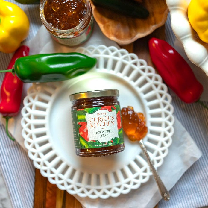 A small jar of hot pepper jelly next to a spoonful of jelly and fresh peppers on a decorative white plate.