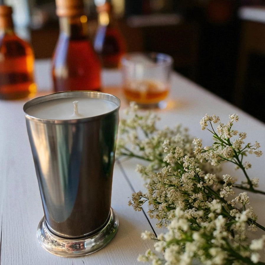 Candle in a metal container on a table with flowers and blurred bottles in the background