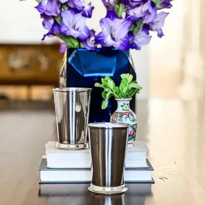 Two silver tumblers on books with a blue vase and purple flowers in the background.