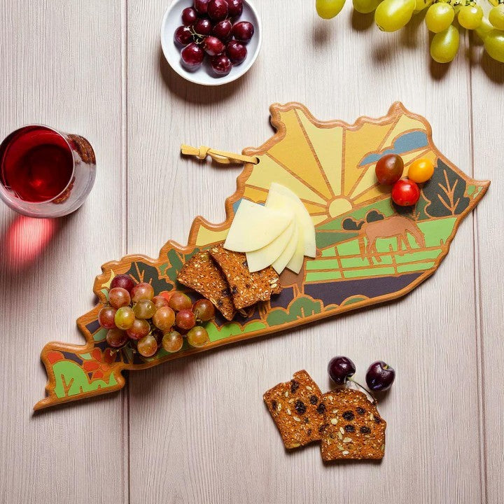 Wooden cutting board with a scenic design, cheese, crackers, and grapes on a wooden surface.