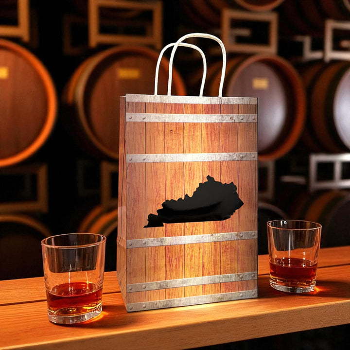 Wooden gift bag with Kentucky outline design on a wooden table with glasses of amber liquid, barrels in the background.