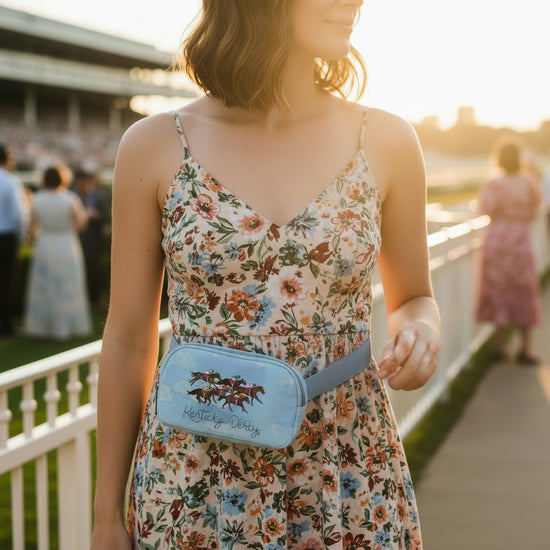 A woman walks through a crowd at Churchill Downs, wearing the Art of the Derby belt bag, with the racetrack and grandstand visible in the background.