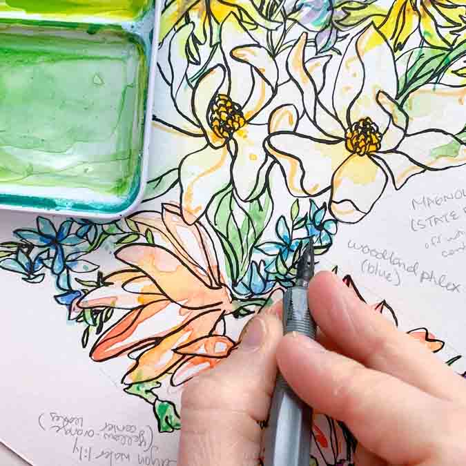 Coloring book with floral designs and a hand holding a pen near a green watercolor pan.