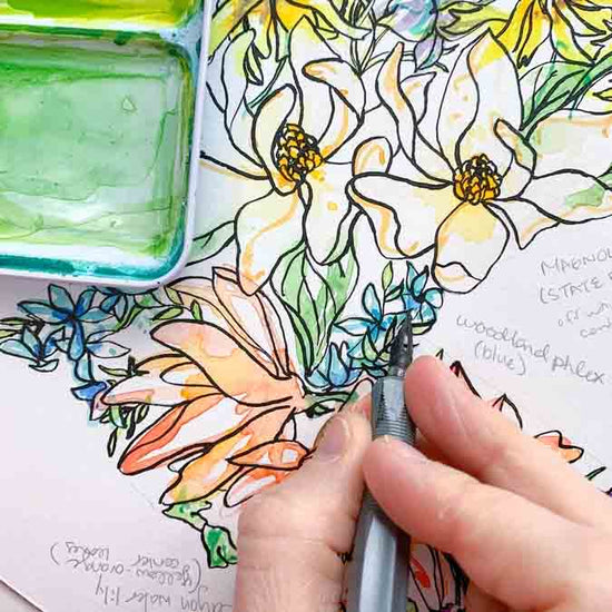 Coloring book with floral designs and a hand holding a pen near a green watercolor pan.