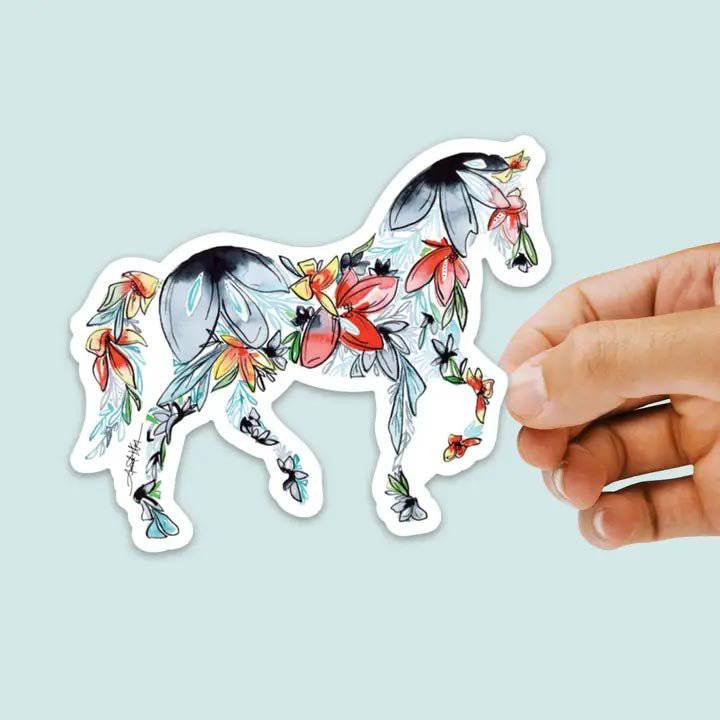 Hand holding a floral-patterned horse sticker against a light blue background