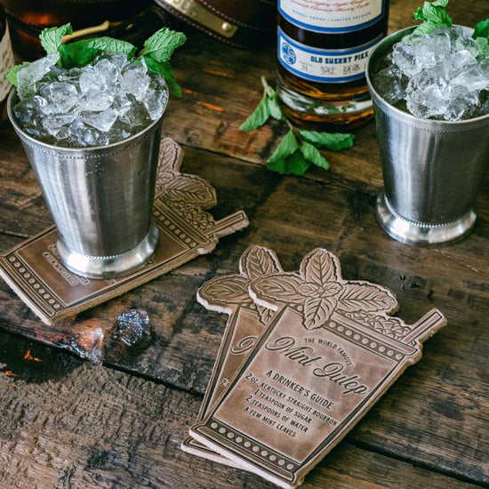 Two silver mint julep cups with ice and mint leaves on a wooden surface, accompanied by a leather coaster titled 'The World Famous Mint Julep'.