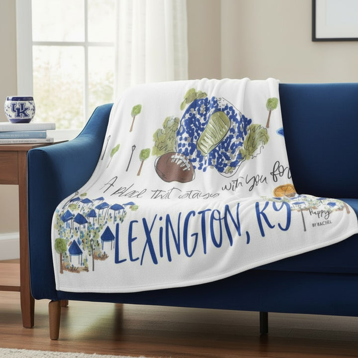 White throw blanket draped over a blue sofa, showing Lexington, Kentucky-inspired watercolor illustrations and "LEXINGTON, KY" text.