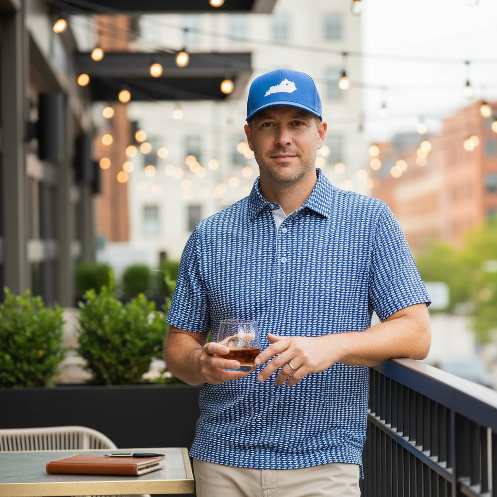 A man wearing the Lexington, Kentucky Performance Polo and a blue hat, smiling while holding a bourbon glass on an urban patio.